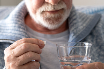 An elderly man with flu or COVID symptoms takes a pill with a glass of water. A stressed, bearded older man takes antidepressants or sedatives. Medications at work.