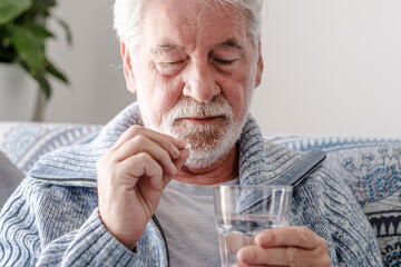 An elderly man with flu or COVID symptoms takes a pill with a glass of water. A stressed, bearded older man takes antidepressants or sedatives. Medications at work.