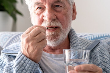 An elderly man with flu or COVID symptoms takes a pill with a glass of water. A stressed, bearded older man takes antidepressants or sedatives. Medications at work.