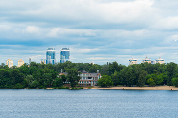 Fototapeta premium Scenic view of a wide river with a sandy beach, lush green trees and modern skyscrapers in the background under a dramatic cloudy sky