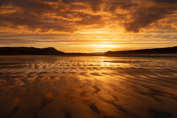 Lever de soleil dor&eacute; sur la plage de l&rsquo;Aber, Finist&egrave;re, Bretagne. Ciel charg&eacute; et reflets sur le sable mouill&eacute; &agrave; mar&eacute;e basse, paysage marin calme.