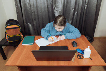 Focused young girl in blue hoodie sitting at wooden desk, writing in notebook and using laptop for online education. Remote learning concept at home