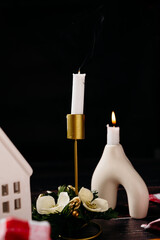 Minimal winter still life with white candle, ceramic house and dark background. Cozy Scandinavian holiday decor and calm festive atmosphere.