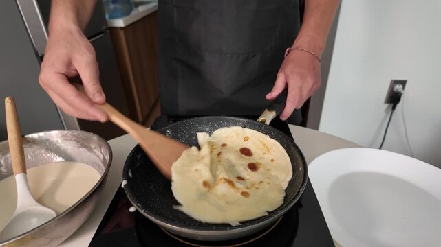 A man in an apron frying pancakes. Close-up. Flips a pancake with a wooden spatula on an induction hob