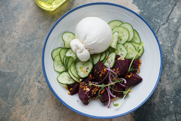 Salad with burrata, cucumber and beets in a white and blue plate, horizontal shot on a beige and grey granite background, elevated view