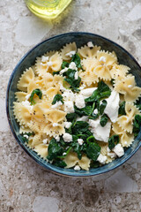 Bowl of farfalle pasta with feta cheese and wilted spinach, vertical shot on a light-brown granite background, elevated view
