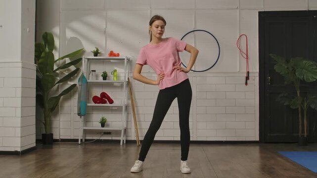 A woman in sportswear performs a pelvic rotation exercise to warm up her muscles, before training on a blue mat in a gym with various equipment. Exercise, fitness training for women, healthy lifestyle