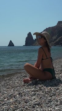 Woman beach ocean, happy woman sitting on pebble beach by the sea, sunny day, travel, vacation. Vertical video
