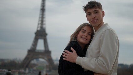 A young couple embraces in Paris France with the Eiffel Tower in the background on an overcast day....