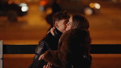 A romantic young couple embraces on a bench in Paris France at night with city lights blurred in...
