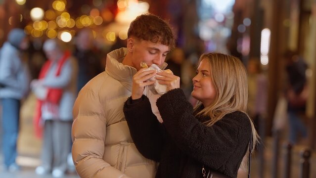 A romantic couple shares a street food snack in Paris France at night The urban city background is blurred with bokeh lights creating a warm and inviting atmosphere for love and travel concepts.