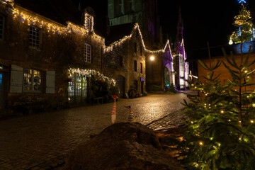 Rue du village de Locronan en Bretagne illumin&eacute;e pour No&euml;l, fa&ccedil;ades en pierre et &eacute;glise en arri&egrave;re-plan, d&eacute;corations lumineuses et ambiance nocturne chaleureuse.