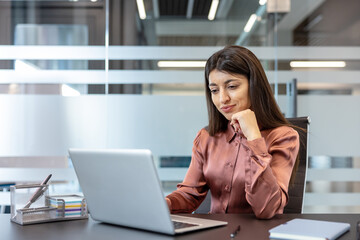 Young businesswoman concentrates on her laptop, making strategic decisions and managing projects in a modern office, projecting confidence, focus, and professional leadership