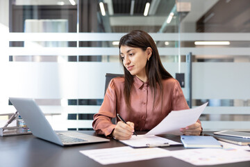 Businesswoman at office desk reviewing documents and taking notes on a clipboard while working on a...