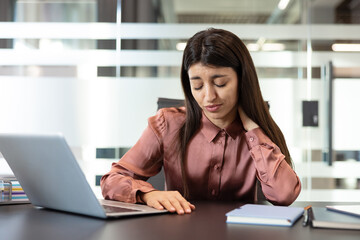 Young businesswoman with dark hair and a pink shirt rubbing her aching neck while sitting at a desk with a laptop, experiencing stress and discomfort during a long workday in the office