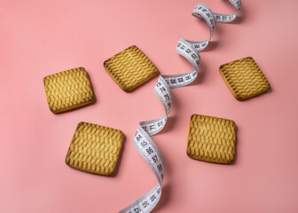 Square cookies are scattered on a pink background with a measuring tape arranged in a curved shape. The composition symbolizes diet, weight loss, calorie control, healthy lifestyle choices, and the ba