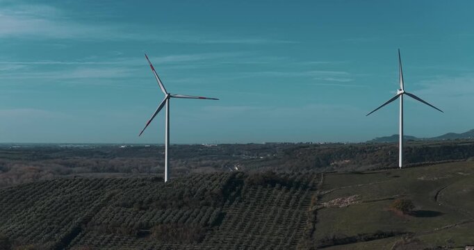 Aerial view of wind turbines standing tall on rolling hills, contrasting with the teal sky and the patterned fields, Serre, Campania, Italy.