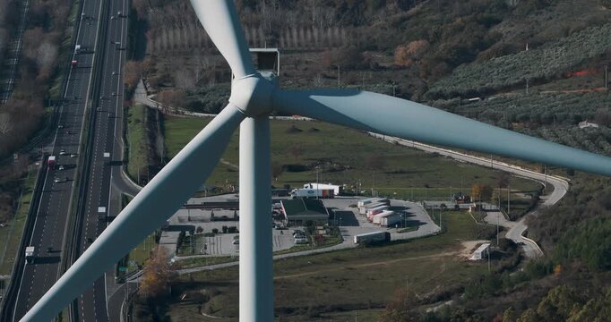 Aerial view of a colossal wind turbine standing tall near a bustling highway amid the undulating hills, Serre, Campania, Italy.