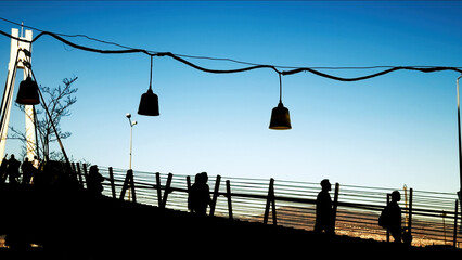 People Walking On Pier With Hanging Lights At Sunset