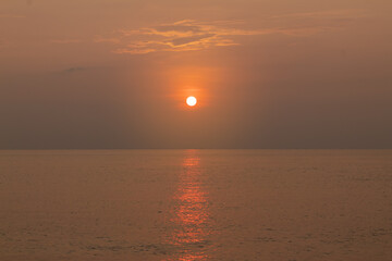 Muted orange sunset over calm ocean water reflecting bright sun disk light path on the surface.