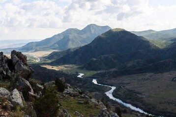 Expansive panoramic view of a winding river meandering through a lush mountain valley with rugged rocky slopes and distant peaks under a bright, cloudy sky.