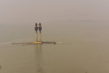 Bridge construction pillars standing in a wide river under hazy sky, infrastructure development concept, calm water reflection, engineering project, and minimal industrial landscape.