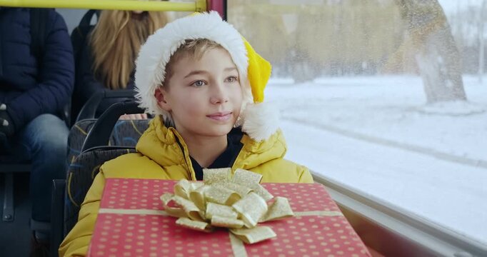 Front view of small boy sitting on bus, smiling. Kid in Christmas hat holding gift box, looking forward, glad, happy. Concept of New Year, Christmas and winter vacations.