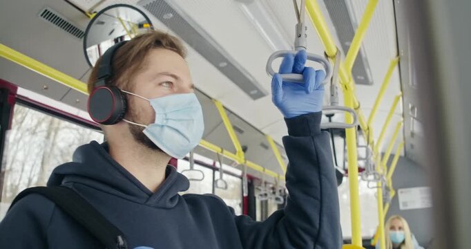 Side view of passengers going by bus, wearing medical masks. man with rucksack and earphones holding grey handle, looking forward Concept of public transport and quarantine.