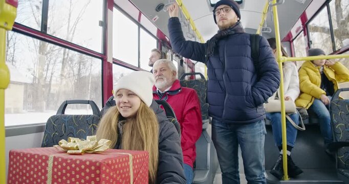 Side view of passengers traveling by bus. Small girl holding gift box, smiling, old man wearing red jacket, sitting, boy standing, handle holding. Concept of public transport.