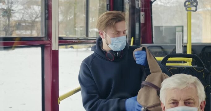 Front view of boy in mask and gloves sitting on bus. Passengers on bus, boy with earphones and rucksack, using smartphone, texting, putting in pocket. Concept of public transport.