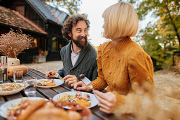 A family gathers around a wooden table outside, enjoying a warm meal on an autumn weekend, surrounded by trees with colorful leaves. Laughter fills the air as they share stories.
