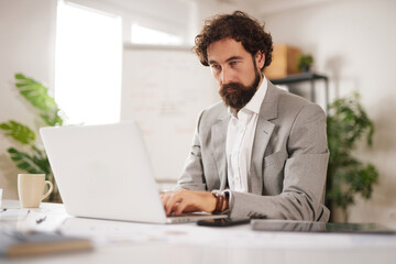 Fototapeta premium A man is sitting in an office chair at a desk. He is focused on his laptop while working. A cup is nearby and there are plants in the background.