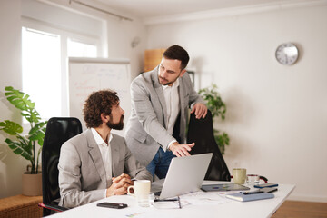 Two team members discuss their project in a light office. One person sits while the other stands, pointing at a laptop. They focus on brainstorming ideas for success.