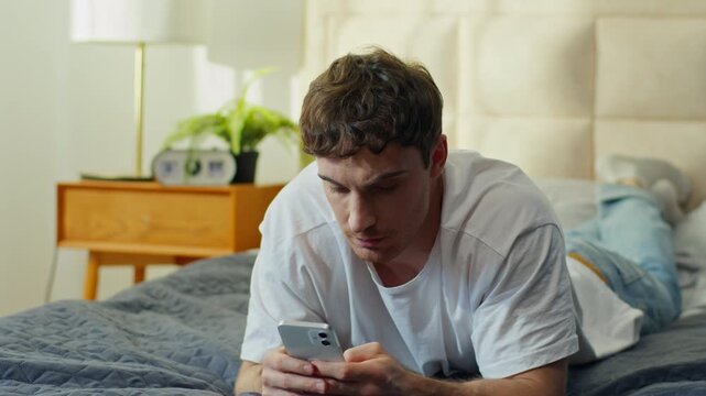 Unhappy Caucasian man lying in bed using smartphone at home worried confused male checking phone problem guy frowning mobile banking fraud spam crash app social media error lost connection breakdown