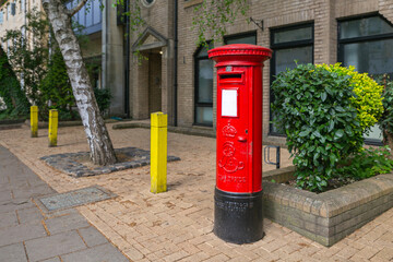 Bright Red Postbox on City Sidewalk with Yellow Bollards and Greenery, Urban Scene