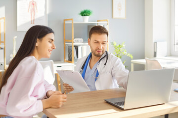 Fototapeta premium Focused doctor in lab coat showing medical results on laptop to smiling patient in clinic. Female patient listening attentively as doctor explaining diagnosis and treatment options during consultation