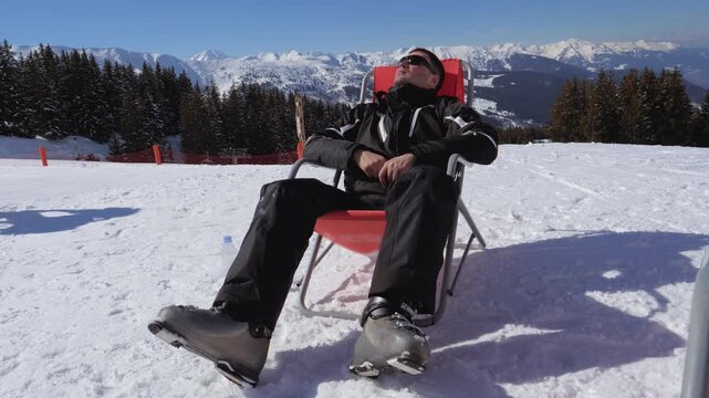 Man sunbathing relaxing in chair on snow mountain. Sunlit alpine resort. Skier wearing ski jacket boots and helmet goggles on forehead. Leisure after lunch during winter vacation