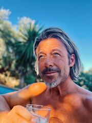 Happy blond man with a stubble beard holding a glass by a pool in a sunny summer garden.