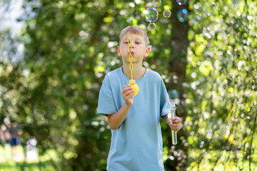 Blond teen boy blowing soap bubbles in summer park on sunny day