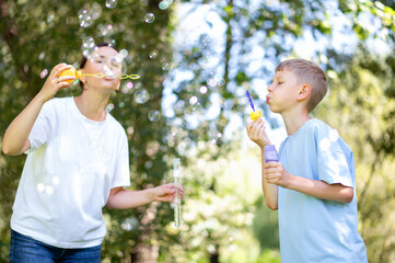 Mother and teen boy having fun blowing soap bubbles in summer park