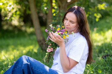 Beautiful woman with long hair sitting on green lawn holding a bouquet of wildflowers outdoors