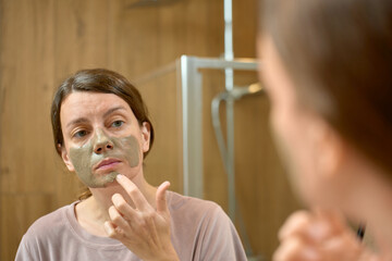 Woman applies clay face mask on face while looking at bathroom mirror