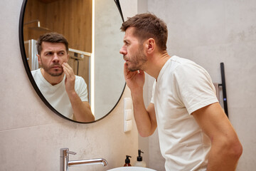 Man examines facial skin while looking at mirror in modern bathroom