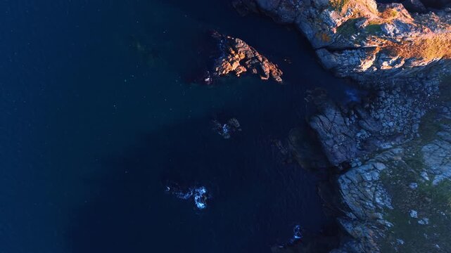Waves crash against the rocky shoreline as the sun sets over the water. Dark tones of the sea contrast with the light on the rocks. Birds can be seen flying in the distance.