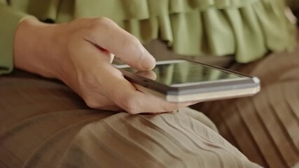 A woman's hand in a green shirt scrolling on a smartphone screen with her thumb over her brown pleated skirt lap - Powered by Adobe