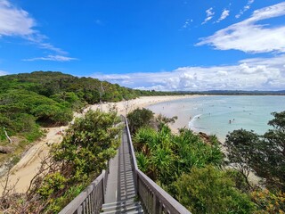 Obraz premium Byron Bay, New South Wales, Australia - 25 December 2025: Beach View. The beach curves gently along the coastline, bordered by lush green vegetation and forested hills. The water is clear turquoise.