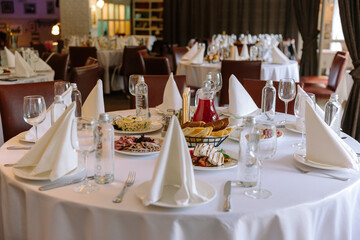 Round banquet tables set with white tablecloths, snacks, appetizers, and drinks in a restaurant for...