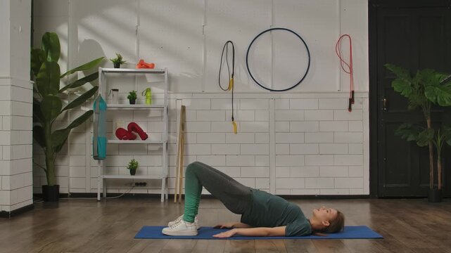 Side view of a woman in sportswear performing a pelvic lift exercise on a blue mat in a gym with various equipment.