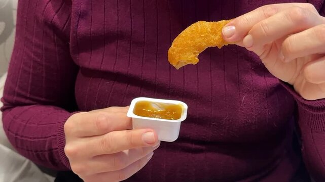 Woman Hand Holding Chicken Nugget Over Sauce, CloseUp Of Casual Eater In Maroon Shirt Dipping Golden Crispy Nugget Into HoneyMustard. Remote Worker Enjoying Quick food.