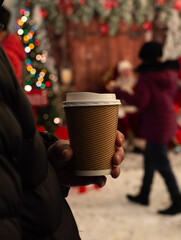 Person Holding Hot Coffee at Christmas Market. Cozy Winter Scene with Coffee Cup and Festive Background
